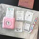 PeriPage Pocket Printer in pink with printed lists on marble surface, featuring cute designs and colorful pen.
