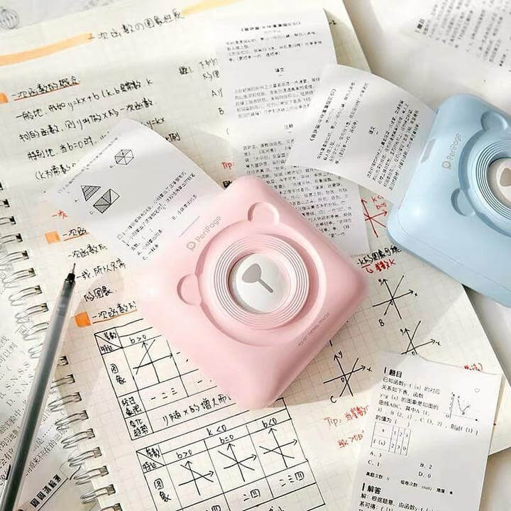 Pink and blue PeriPage Pocket Printer printing notes on a desk with an open notebook and pen nearby.